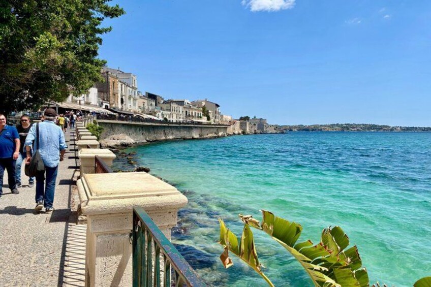 Une promenade en bord de mer où l’on voit des gens se promener, une eau turquoise et limpide, et des bâtiments historiques en arrière-plan. 5