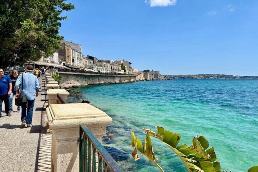 A coastal walkway with people walking, clear turquoise water, and historic buildings in the background. 5