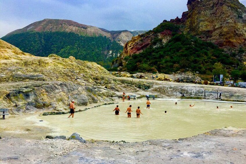 A geothermal spa with people bathing in mineral-rich waters, surrounded by rocky terrain and mountains. 3