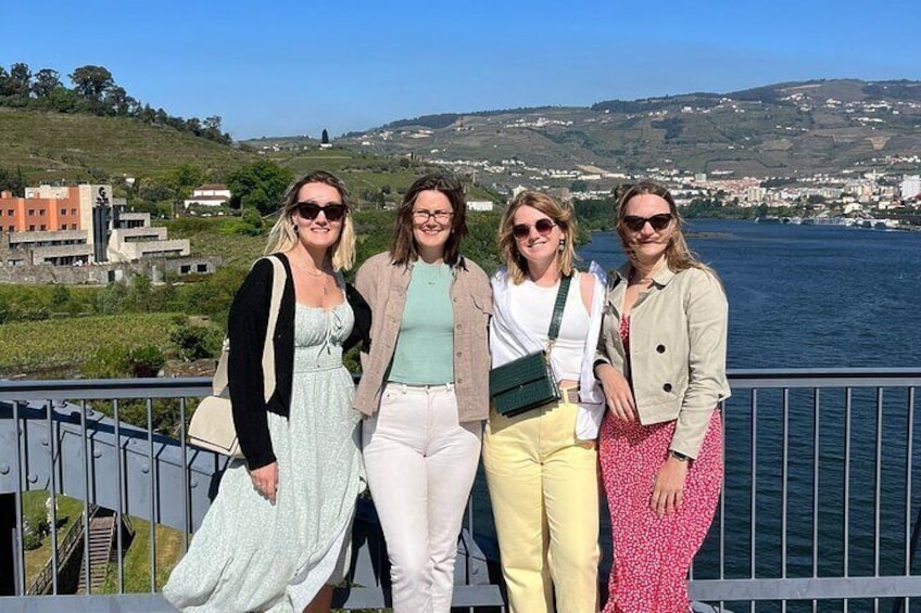 Quatre personnes se tiennent sur un pont, d’où l’on peut admirer un paysage pittoresque composé d’une étendue d’eau et de collines en arrière-plan. 3