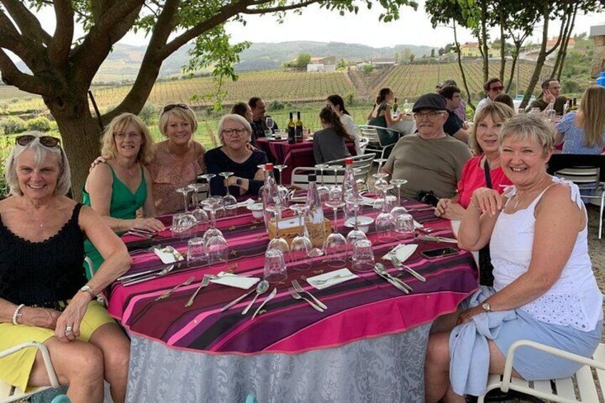 A group of people seated at an outdoor table with a tablecloth, surrounded by a vineyard landscape. 2