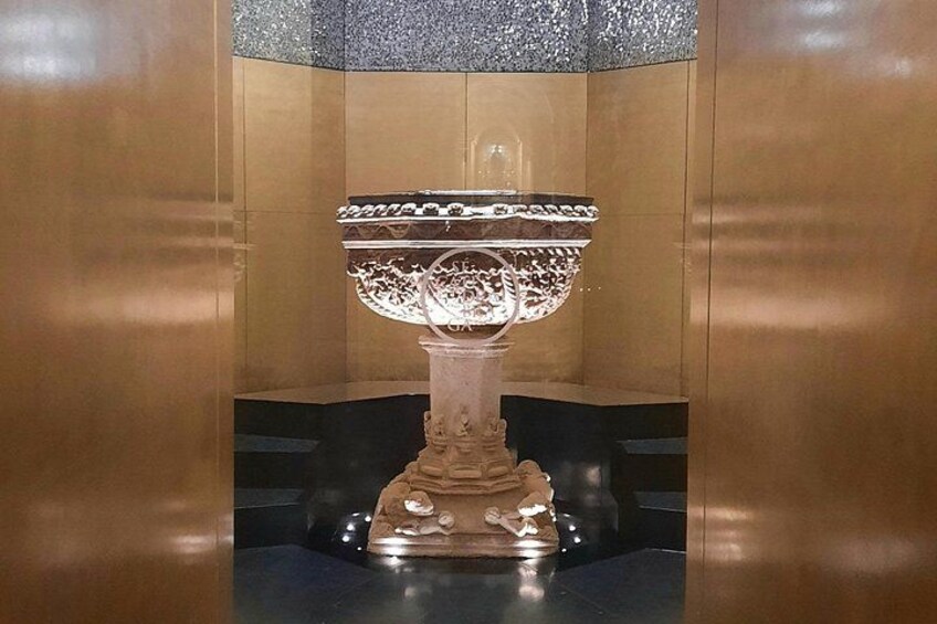 A decorative urn with intricate designs, placed on a pedestal against a wooden wall. 2