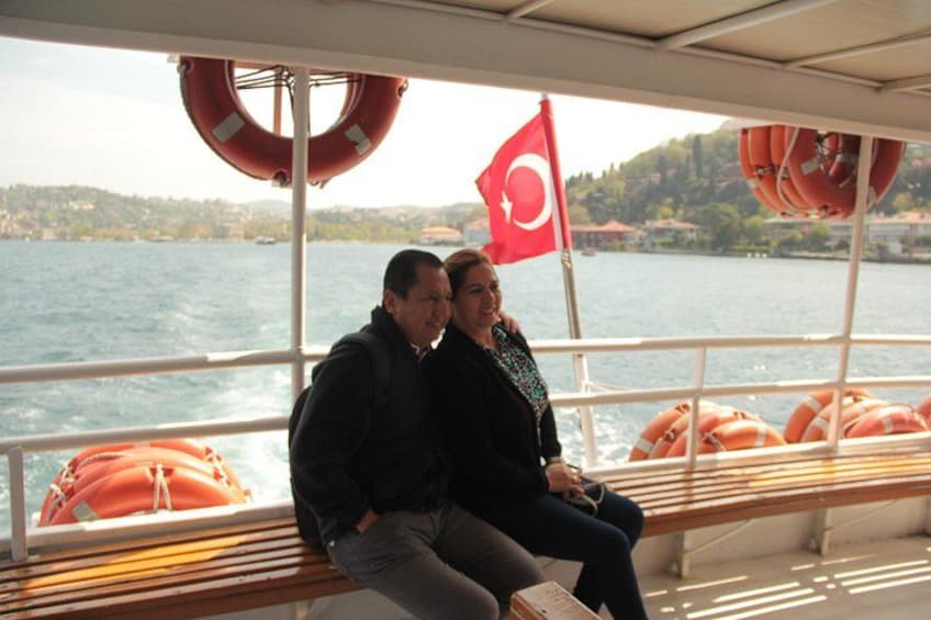 Two people sitting on a bench on a boat with lifebuoys and a flag in the background. 1