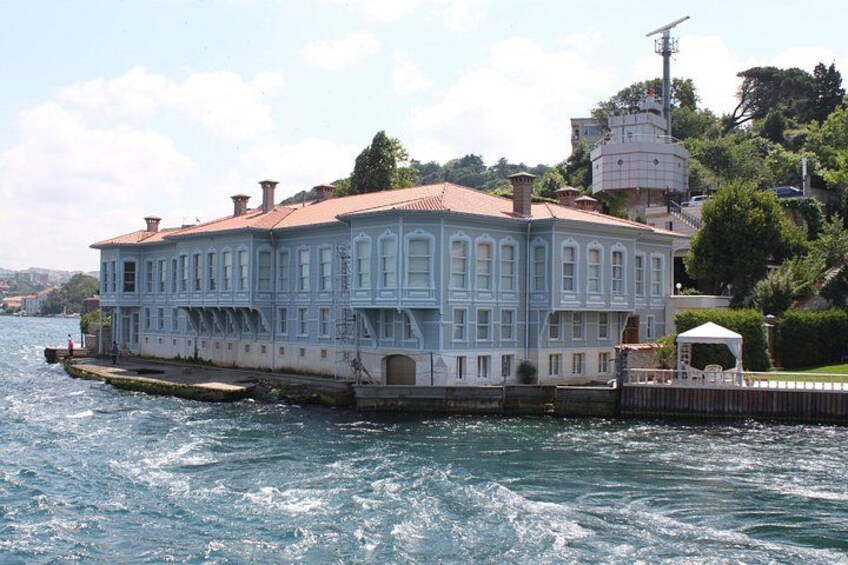 A large, light blue building with a red roof, situated on a waterfront with a dock extending into the water. 2
