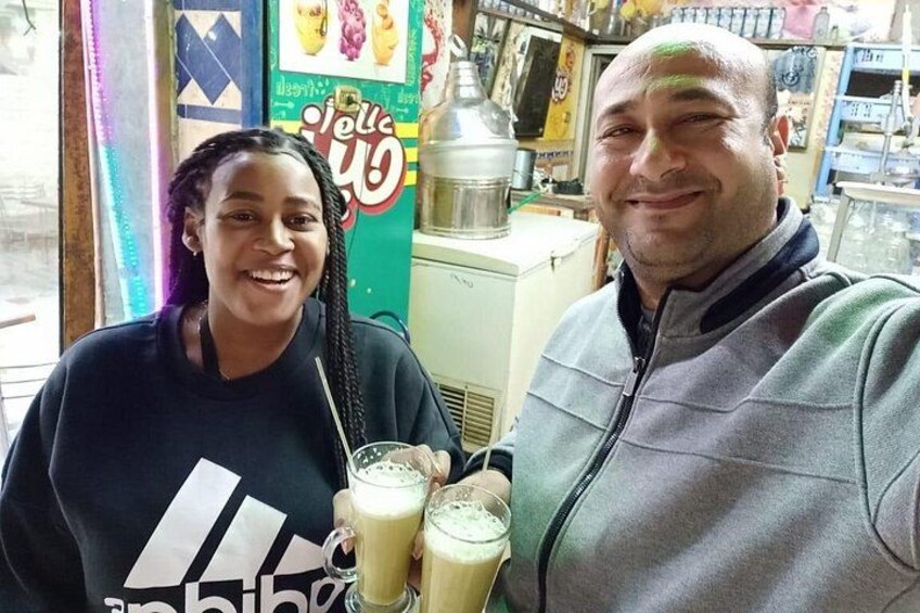 Twee personen met drankjes in een café, met een kleurrijk bord en een koffieapparaat op de achtergrond. 2