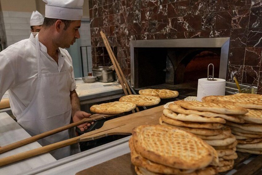 Een kok in een wit uniform bereidt platbroodjes op een houten plank, met een marmeren muur en een bakoven op de achtergrond. 3