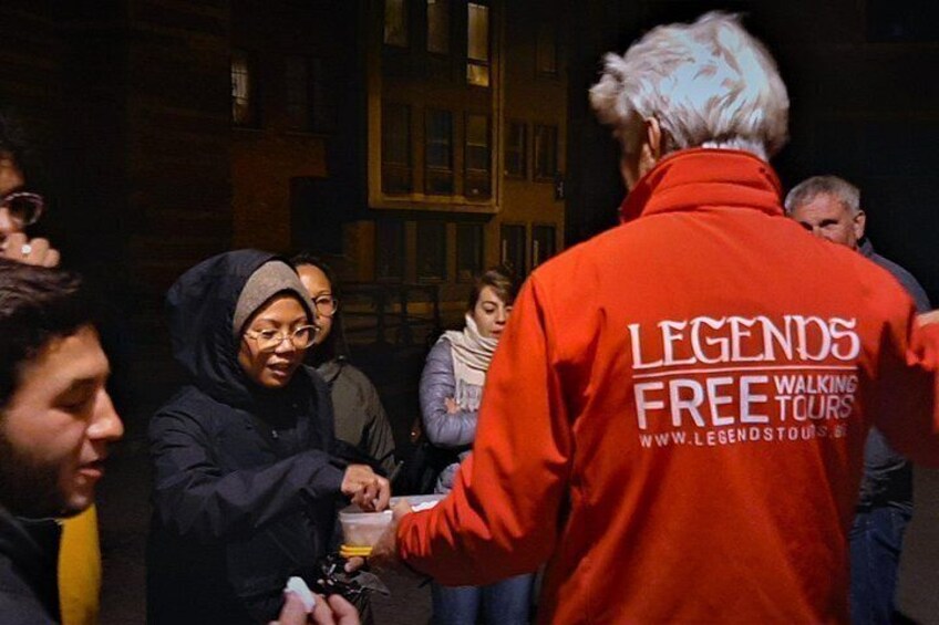 En gruppe mennesker samlet omkring et bord, en person i en orange jakke med teksten 'LEGENDS FREE WALKING TOURS', og en bygning i baggrunden. 4