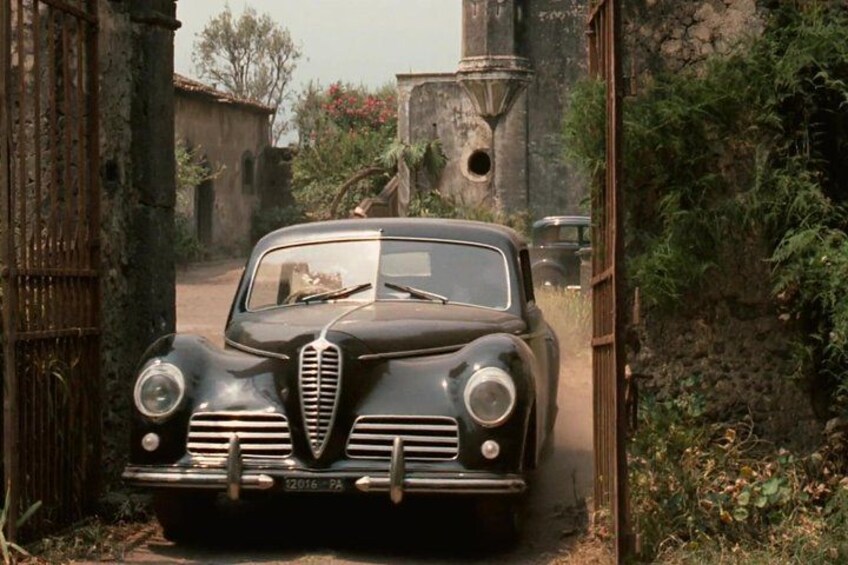 A vintage car parked in a rustic setting with a gate and greenery in the background. 4