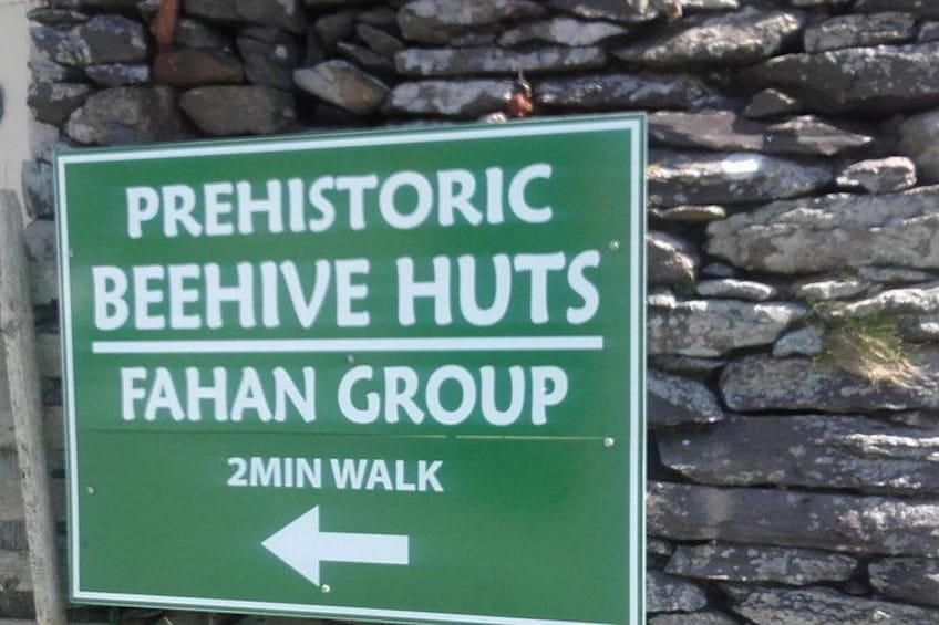 A green sign with white text indicating the direction to 'PREHISTORIC BEEHIVE HUTS' and 'FAHAN GROUP' with a '2MIN WALK' arrow. 1