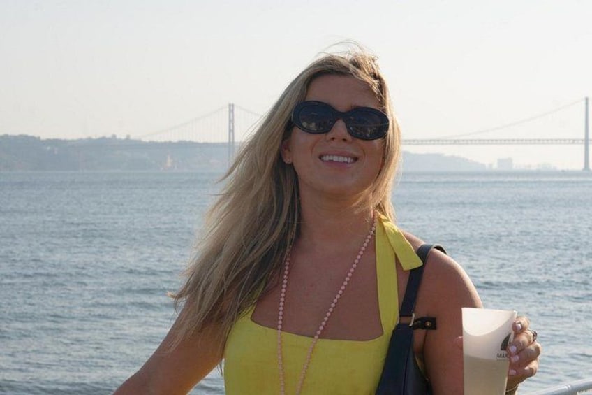 A person wearing a yellow top and a pearl necklace, holding a cup, with a bridge in the background. 2