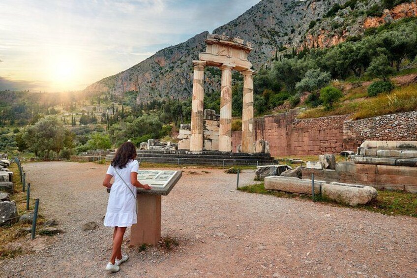 A woman in a white dress standing at a stone pedestal with a tablet, overlooking ancient ruins and a mountainous landscape. 3