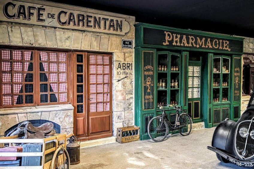 Una fachada antigua con un letrero que dice 'CAFÉ DE CARENTAN' y 'PHARMACIE', con una bicicleta estacionada afuera. 5