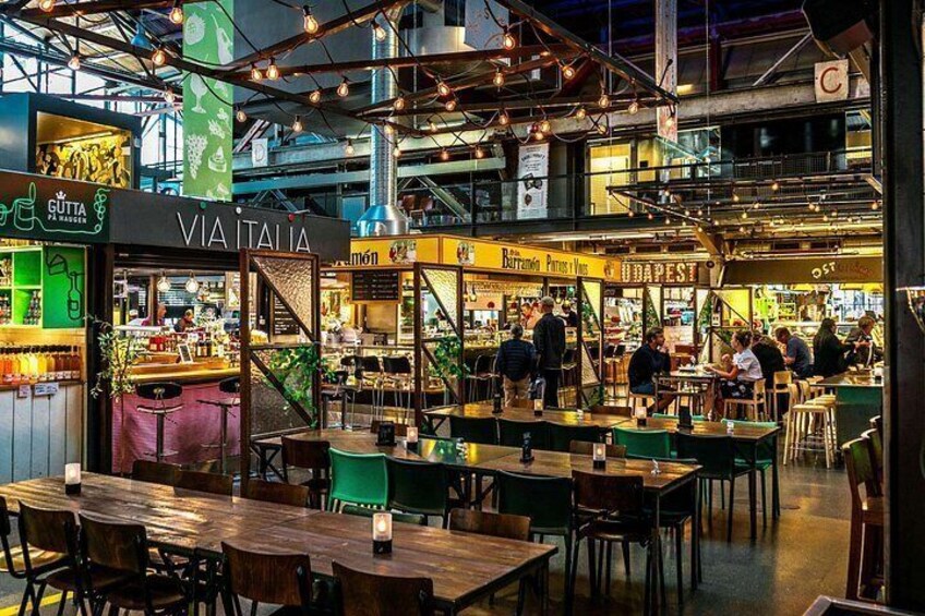 An indoor market with wooden tables and chairs, various food stalls, and hanging lights. 3