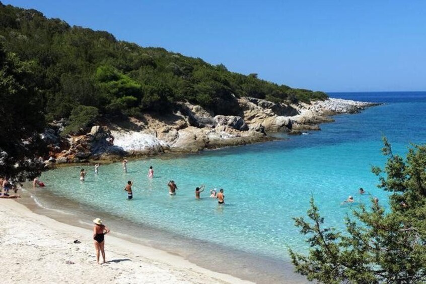 En strand med krystalklart turkis vand, folk der bader, og klippestykker i baggrunden. 4
