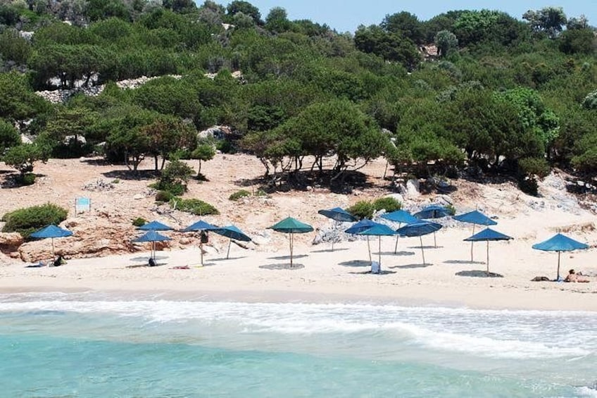 En strand med blå parasoller, klart turkis vand og en skovklædt bakke. 1