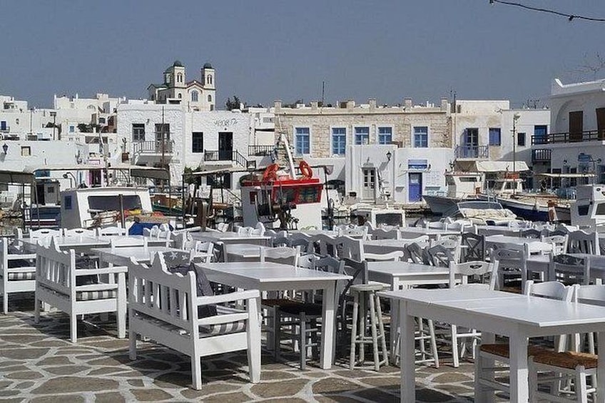 Een café aan zee met witte tafels en stoelen, boten afgemeerd in de buurt, en witte gebouwen met blauwe accenten op de achtergrond. 2