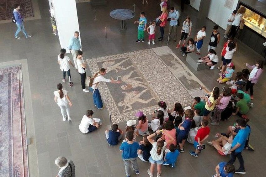 Eine Gruppe von Menschen hat sich um eine große, kunstvolle Fußbodenmosaik in einem geräumigen Innenbereich mit hohen Decken und Säulen versammelt. 2