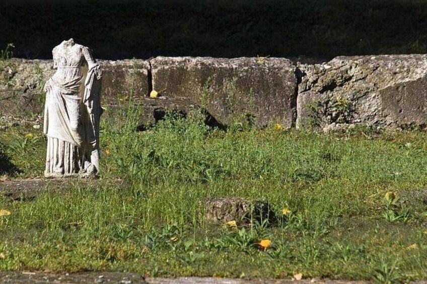 A weathered stone statue in a grassy field with overgrown vegetation and a stone wall in the background. 2