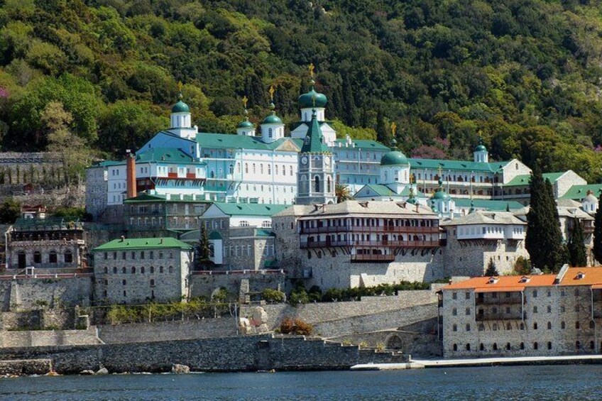 A historic monastery complex with multiple green-domed buildings, a stone wall, and a river in the foreground. 1