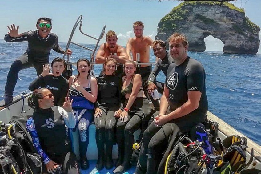 A group of people on a boat with scuba diving equipment, a rocky island in the background, and clear blue water. 1