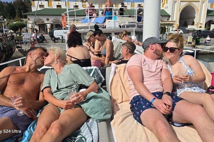 Personas relajándose en un bote, un puerto deportivo con embarcaciones y un edificio con arcos al fondo. 4