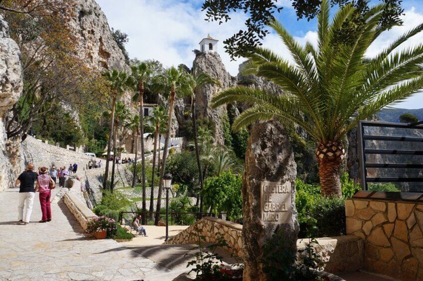 A rocky landscape with palm trees, a stone wall, and a building in the background. 5
