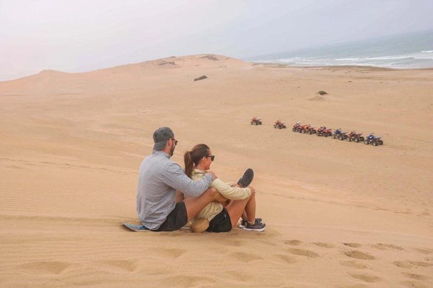 Due persone sedute sulle dune di sabbia, con una fila di ATV sullo sfondo. 3