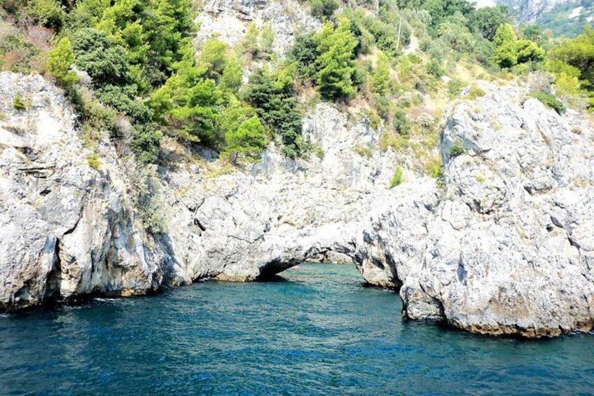 A natural rock arch over a body of water, surrounded by rocky terrain and greenery. 2