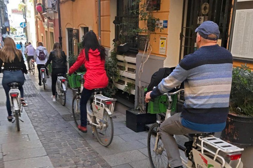 People riding bicycles on a cobblestone street, with buildings and greenery in the background. 3