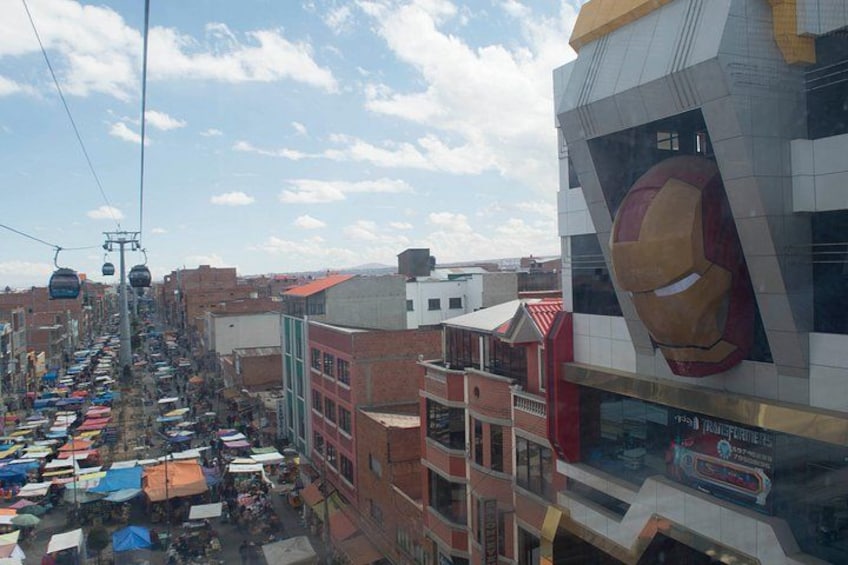 A bustling street with market stalls, a large building with a prominent Iron Man logo, and a cable car system. 2