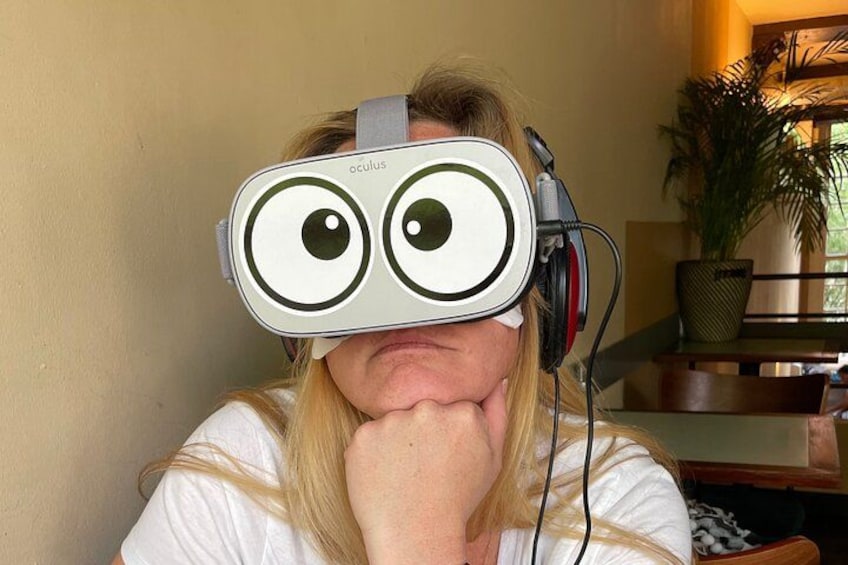 A person wearing a VR headset with a white and green design, sitting in a room with a plant and a wooden table in the background. 2