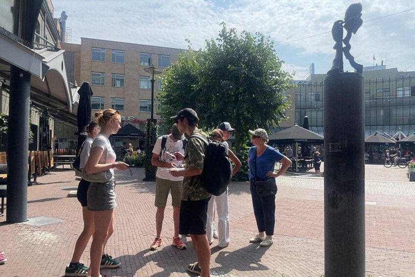 En grupp människor som står på ett gatstenbelagt område i en stadsmiljö, med byggnader och träd i bakgrunden. 4