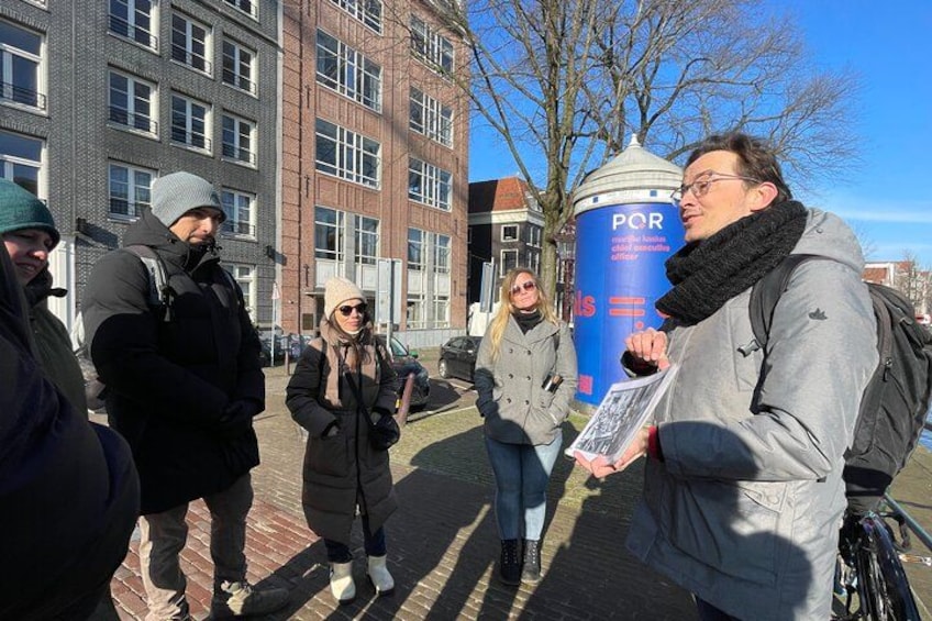 A group of people standing on a city street with buildings and a blue pillar with text. 5