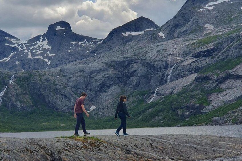 Två personer promenerar längs en stenstrand med ett bergigt landskap i bakgrunden. 2