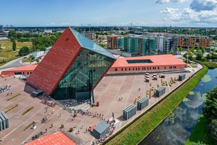 A modern building with a red roof and glass facade, surrounded by a green park and a river. 1