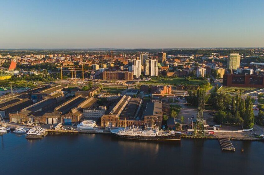 A waterfront cityscape with industrial buildings, a large ship docked at the port, and a clear sky. 2