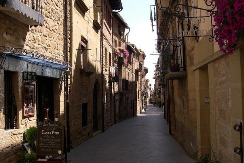 A narrow street lined with historic buildings, a sign for Casa Vino, and balconies with flowers. 2