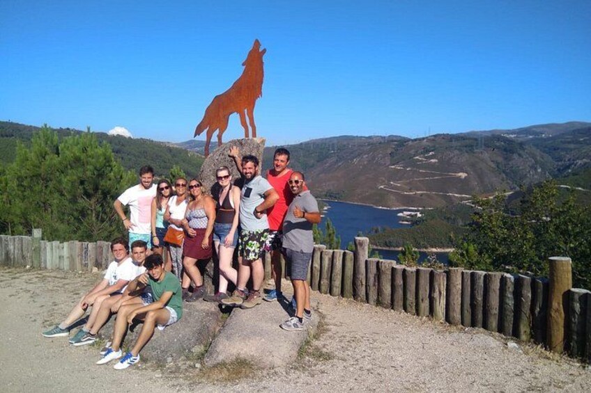 Un groupe de personnes posant devant une statue de loup, sur un belvédère offrant une vue panoramique sur un lac et des collines. 1