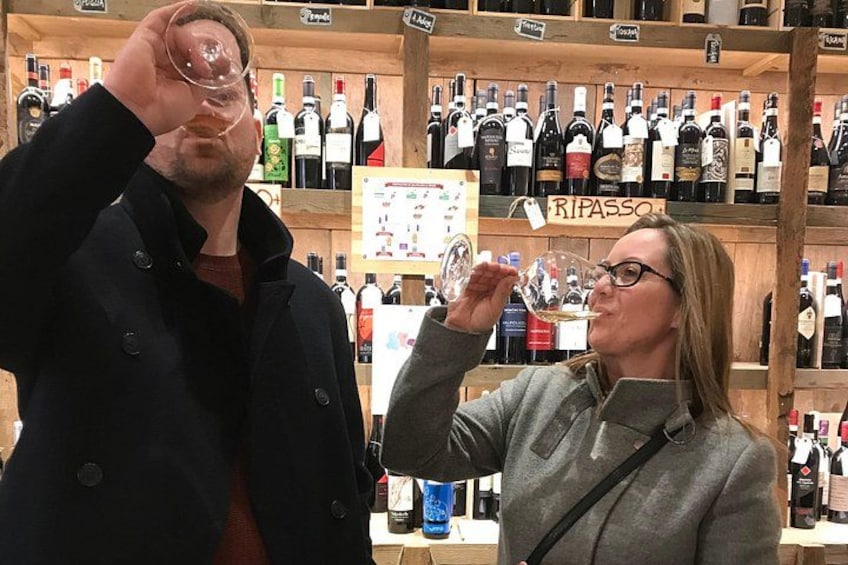 A person holding a wine glass, another person tasting wine, and a shelf with various wine bottles. 3