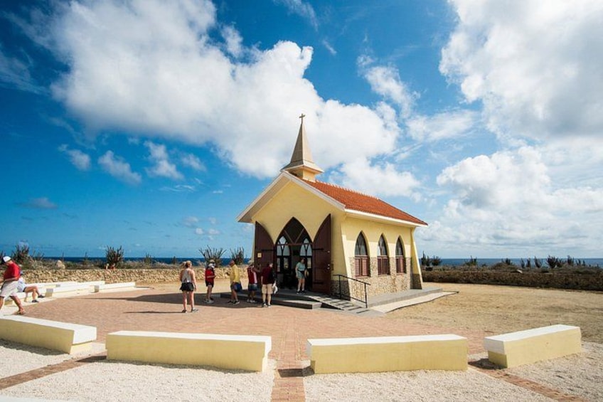 Une petite église avec un toit rouge et des fenêtres en arc de cercle, entourée d’une zone pavée où se trouvent des bancs. 2