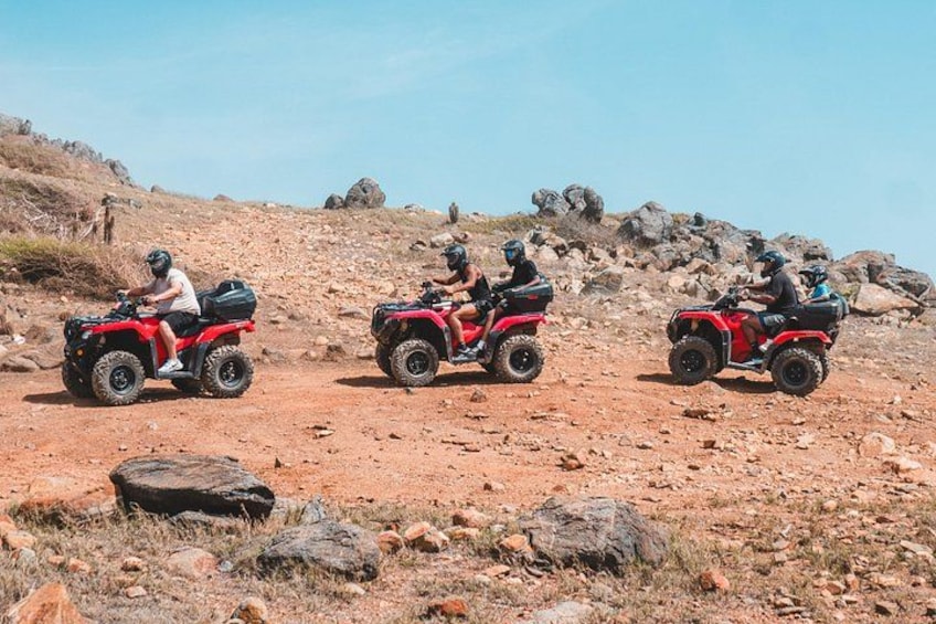 Trois personnes conduisent des quads rouges sur un sentier rocailleux et accidenté, où la végétation est clairsemée. 3