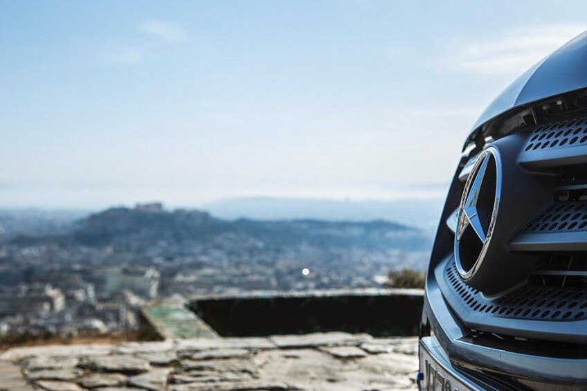 A car's front grille with a logo, a stone ledge, and a cityscape in the background. 5