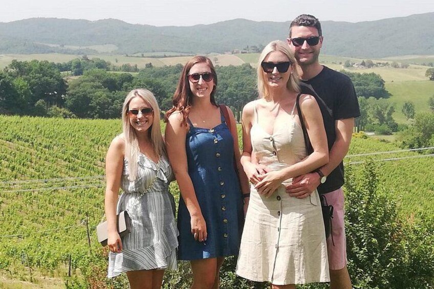 A group of people standing in a vineyard with rows of grapevines and a hilly landscape in the background. 1