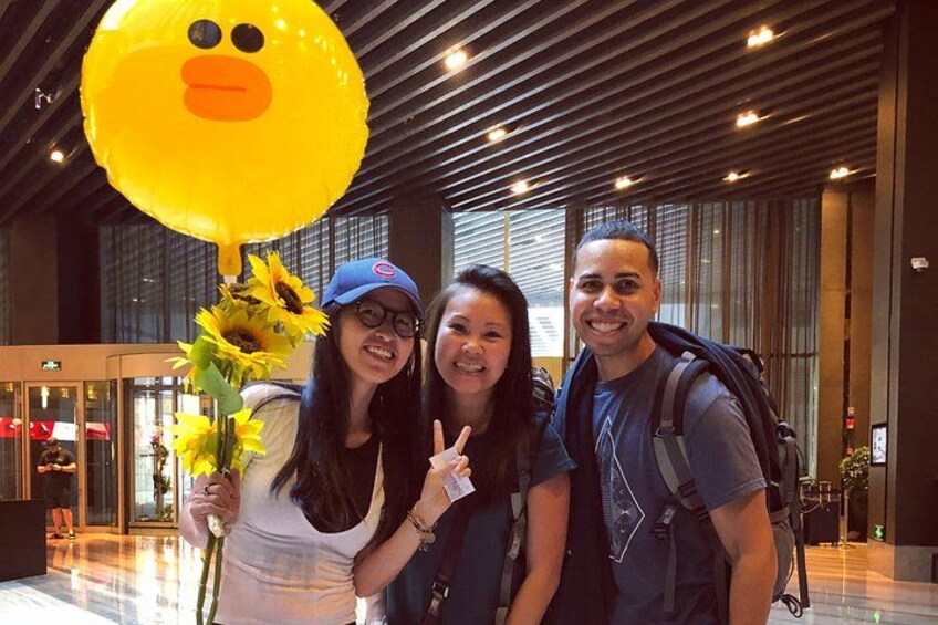 Three people in an indoor setting with a large yellow balloon, a flower, and a person making a peace sign. 2