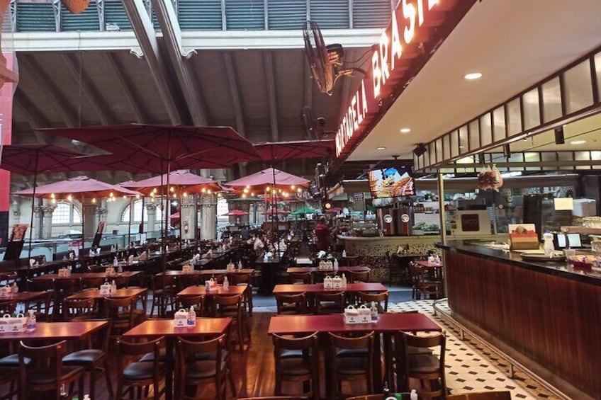 Un restaurant avec des tables en bois et des parasols rouges, un comptoir où sont exposés des produits alimentaires, et une enseigne sur laquelle on peut lire « BRASILIA ». 4