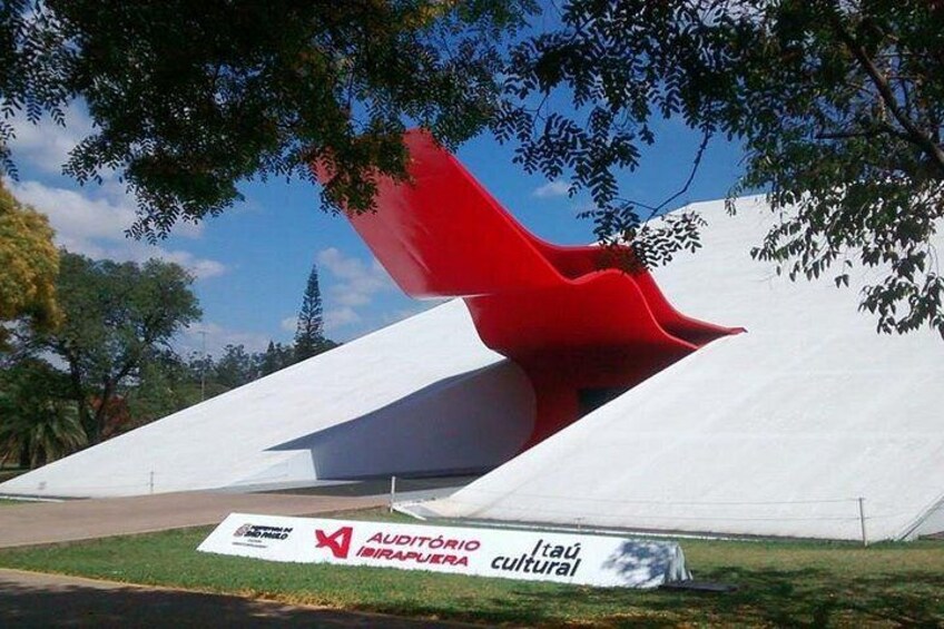 A modern sculpture with a red and white design, located in a grassy area with trees and a clear sky. 1