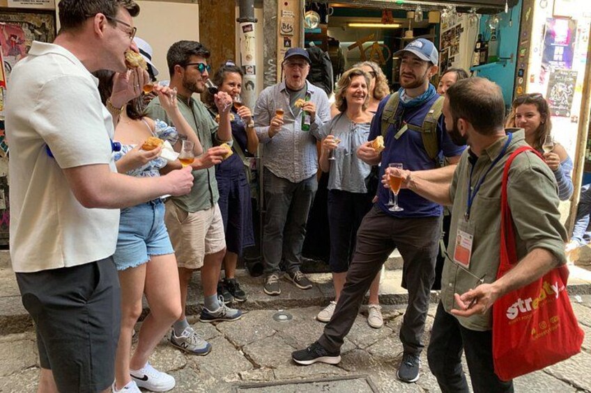 A group of people standing on a street, some holding food and drinks. 2