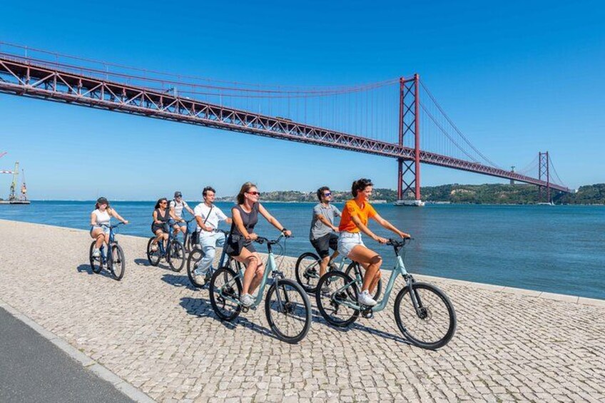 People riding bicycles along a waterfront promenade with a large red bridge in the background. 2