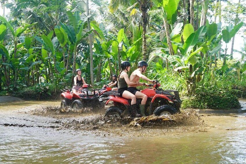 Two people on a red ATV navigating through a muddy water crossing surrounded by dense tropical vegetation. 3