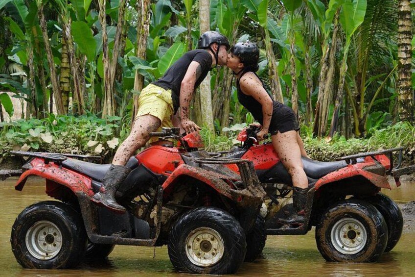 Two people on a red ATV in a muddy area with water and greenery in the background. 3